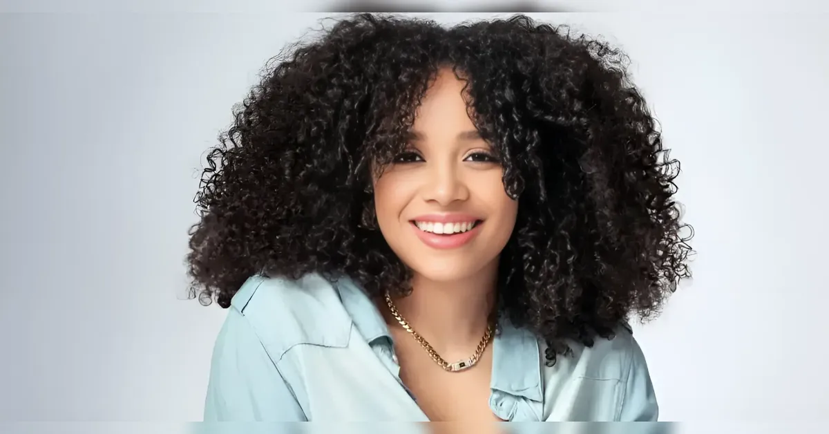 Smiling woman with tight curly hair wearing a light blue shirt and gold necklace, posed against a neutral background.