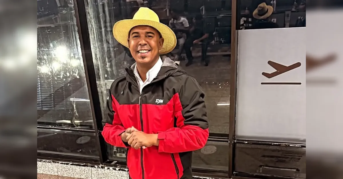 Smiling man wearing a wide-brim straw hat and a red-and-black jacket, standing near glass doors at an airport terminal.