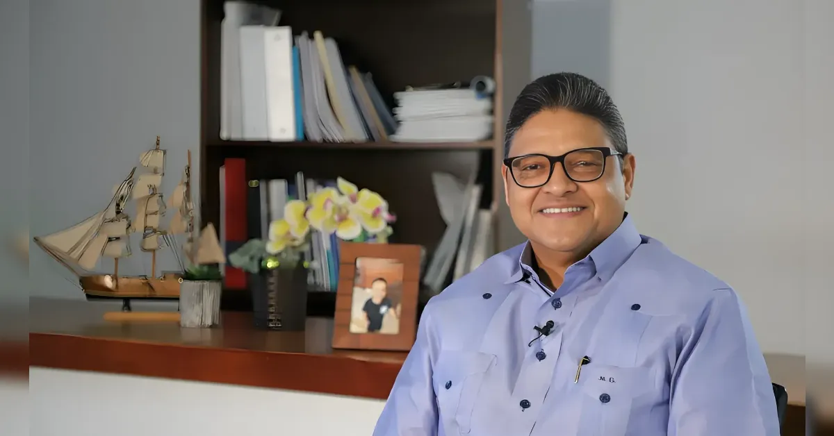 Smiling man with glasses sits at a desk in an office, with a ship model, books, and a photo frame behind him.