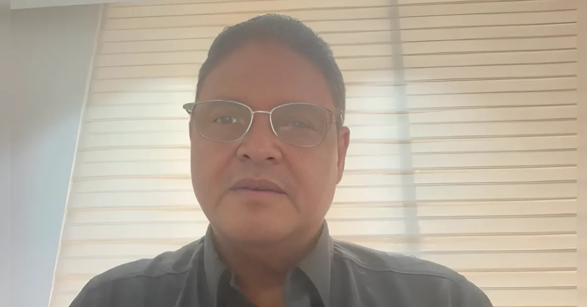 Man wearing glasses looking at the camera, indoors with horizontal blinds behind him.