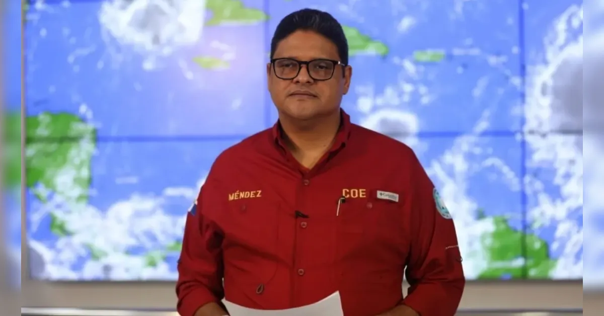 Male presenter in a red shirt stands in front of a large world map display, holding papers for a broadcast.