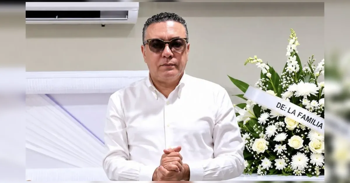 Man in a white shirt and sunglasses stands with hands clasped in front, at a funeral arrangement featuring white flowers and a ribbon reading 'DE: LA FAMILIA'