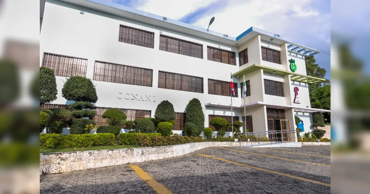 White multi-story building with barred windows, neatly trimmed shrubs, and a paved parking area in front; flagpoles near the entrance.