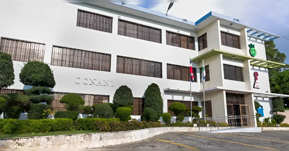 White multi-story institutional building with grid-like windows, surrounded by neatly trimmed shrubs and a small garden area in front.