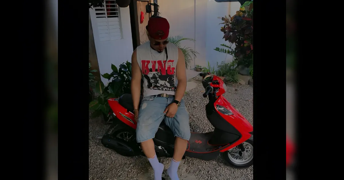 Young man wearing a red cap and sleeveless shirt that says 'KING,' light denim shorts, and lavender socks, sits on a red scooter in a gravel driveway with plants nearby.
