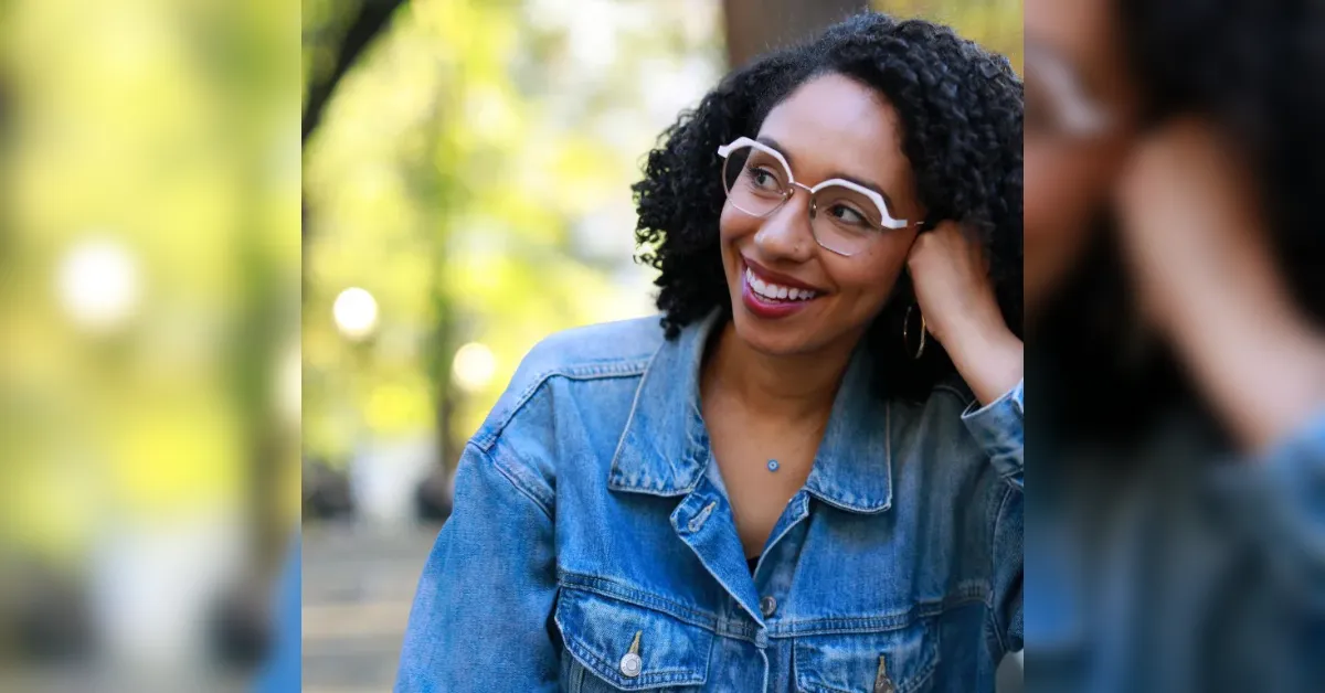 Smiling woman with curly hair and round glasses in a denim jacket outdoors, hand resting on her cheek.