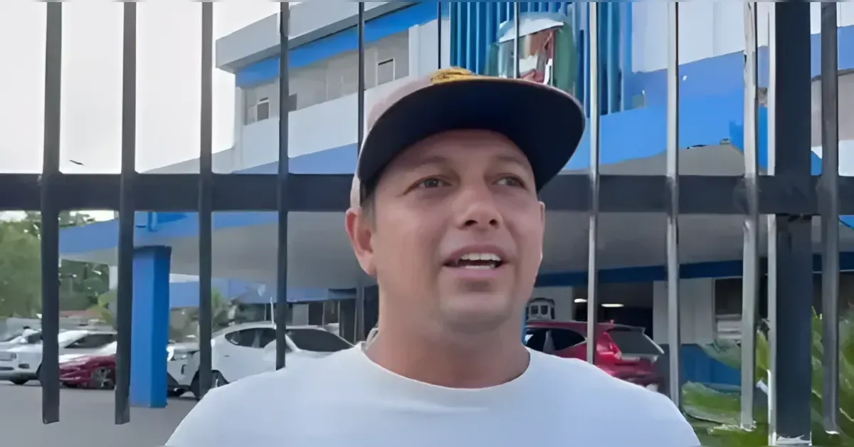 Man in a white shirt and cap speaks to the camera outdoors, with a blue-and-white building and metal fence behind him.