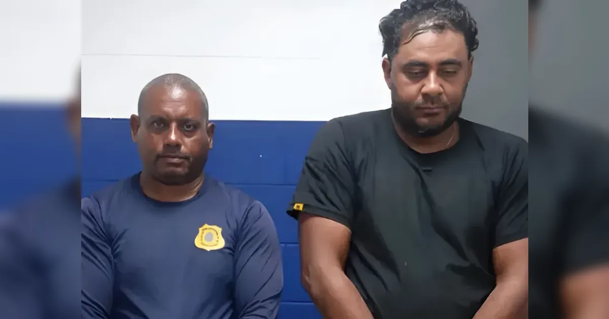 Two men sit side by side against a blue and white wall; the man on the left wears a navy shirt with a yellow badge.