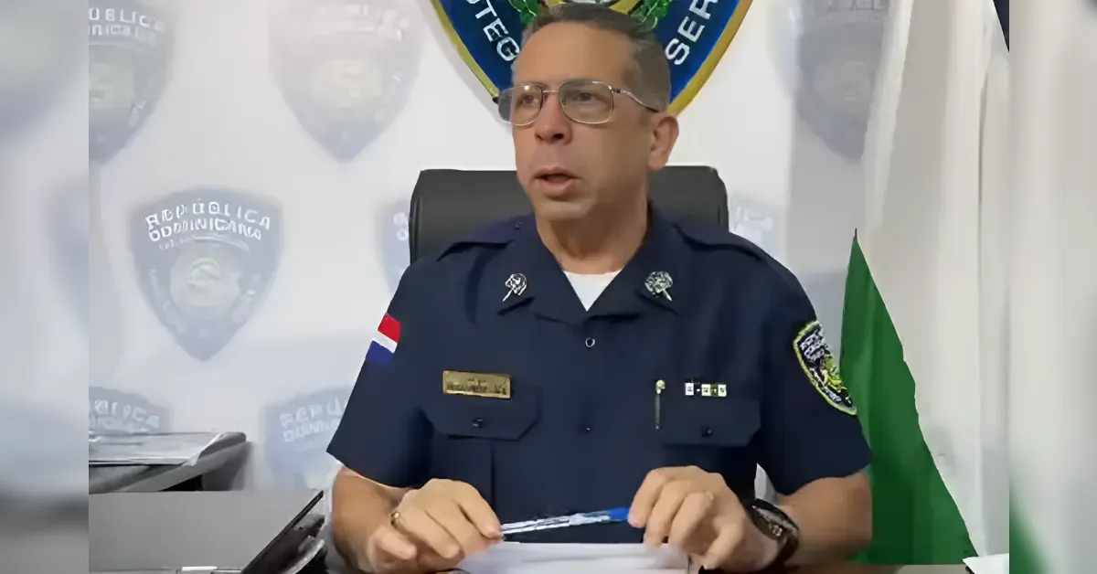 Police officer in a navy uniform seated at a desk with papers, wearing glasses, speaking during a briefing with badges and a flag in the background.