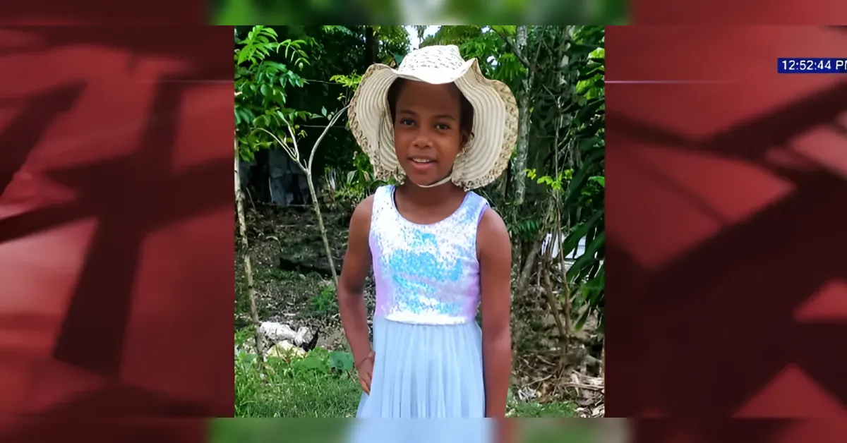 Girl posing outdoors in a light blue dress and wide-brimmed straw hat, smiling at the camera in a garden setting