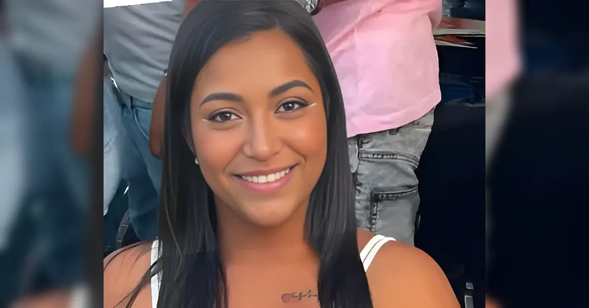 Young woman with straight black hair smiling at the camera, wearing a white tank top and a small tattoo on her collarbone area.