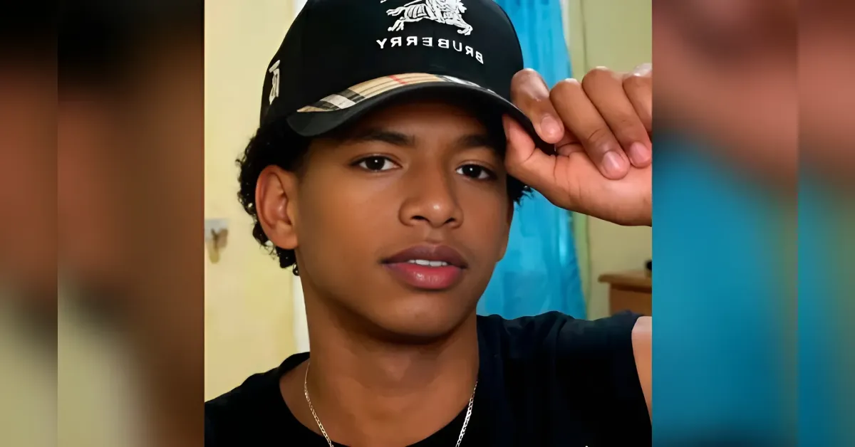 Portrait of a young man indoors wearing a black Burberry cap and a black shirt, touching the brim with his hand.