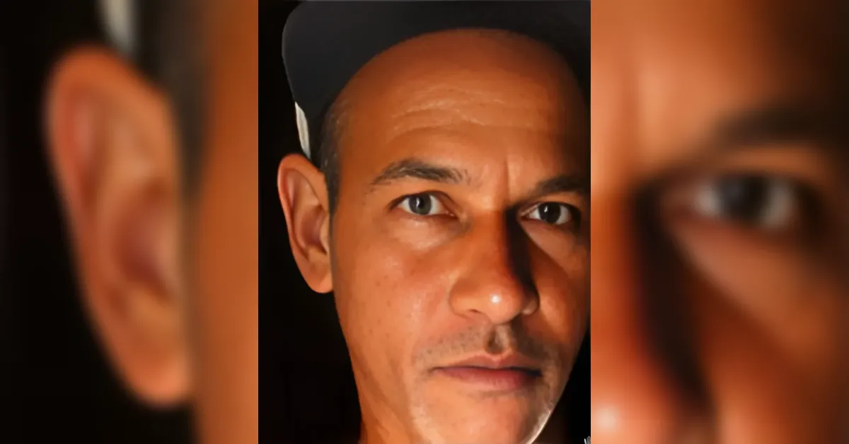 Close-up portrait of a man wearing a dark cap, looking directly at the camera with a neutral expression in low light.