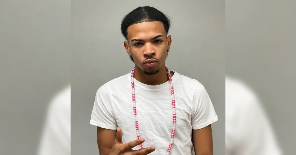 Young man with a white t-shirt and red-and-white beaded necklace looking at the camera against a gray backdrop, making a hand gesture.