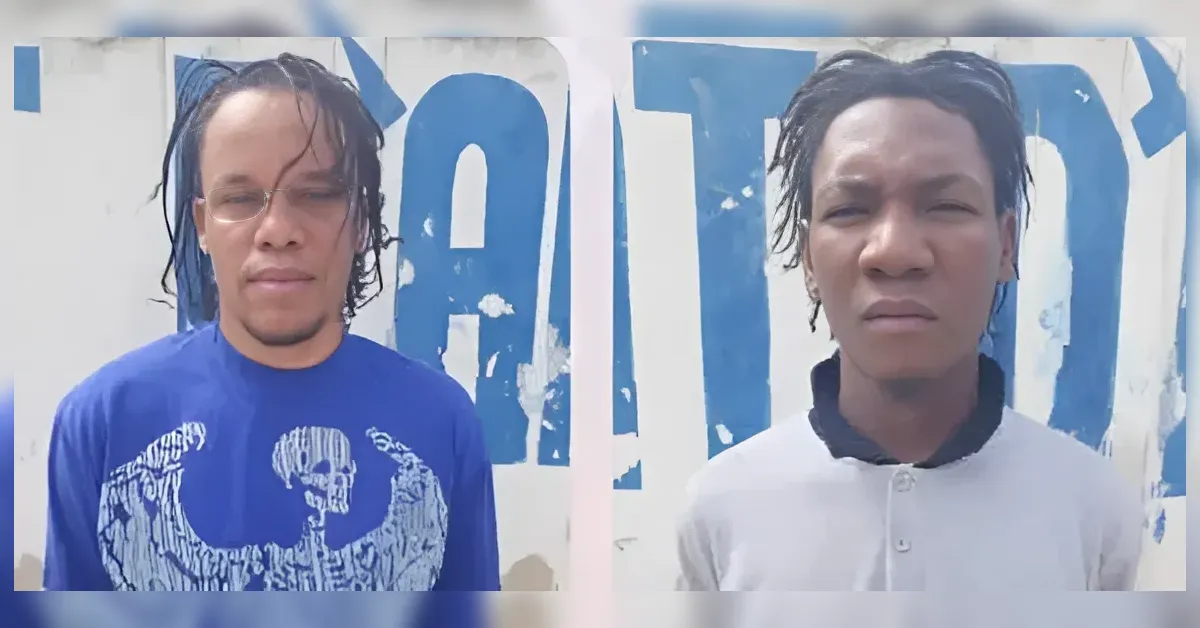 Two men stand side by side in front of a wall with large blue letters; left wears a blue graphic shirt and glasses, right in a white polo with a black collar.