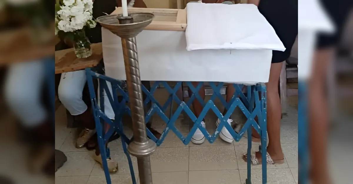 White coffin on a blue portable bier at a funeral, with a tall candle holder in the foreground and flowers in the background.