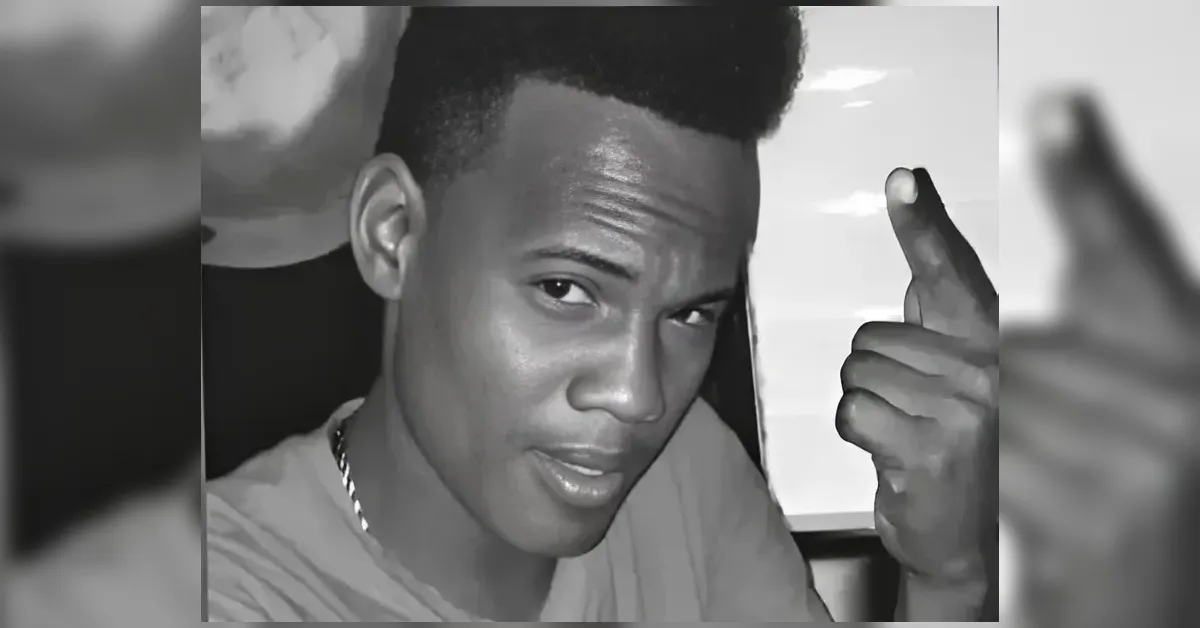 Black-and-white close-up selfie of a young man looking at the camera, making a finger-gun gesture with his right hand.