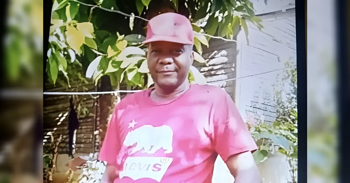 Smiling man in a red cap and red T-shirt with a white graphic, posing outdoors among green leaves.