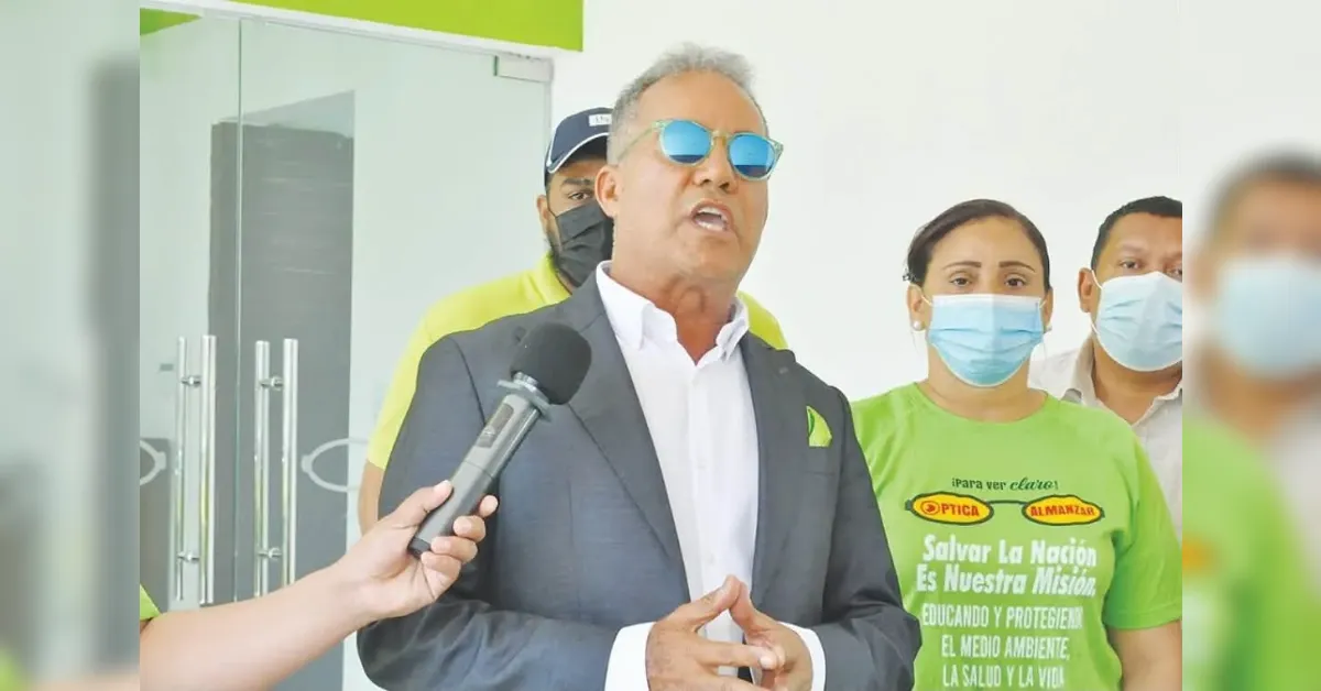 Spokesperson in a suit and blue sunglasses speaks to reporters, microphone held near him, with team members in green shirts behind him.