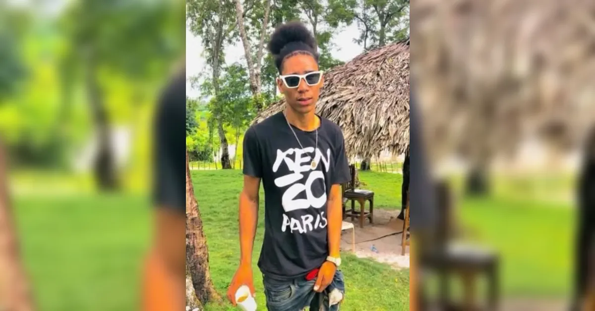 Young man wearing a black Kenzo Paris T‑shirt and white sunglasses stands in a grassy park with a thatched hut in the background.