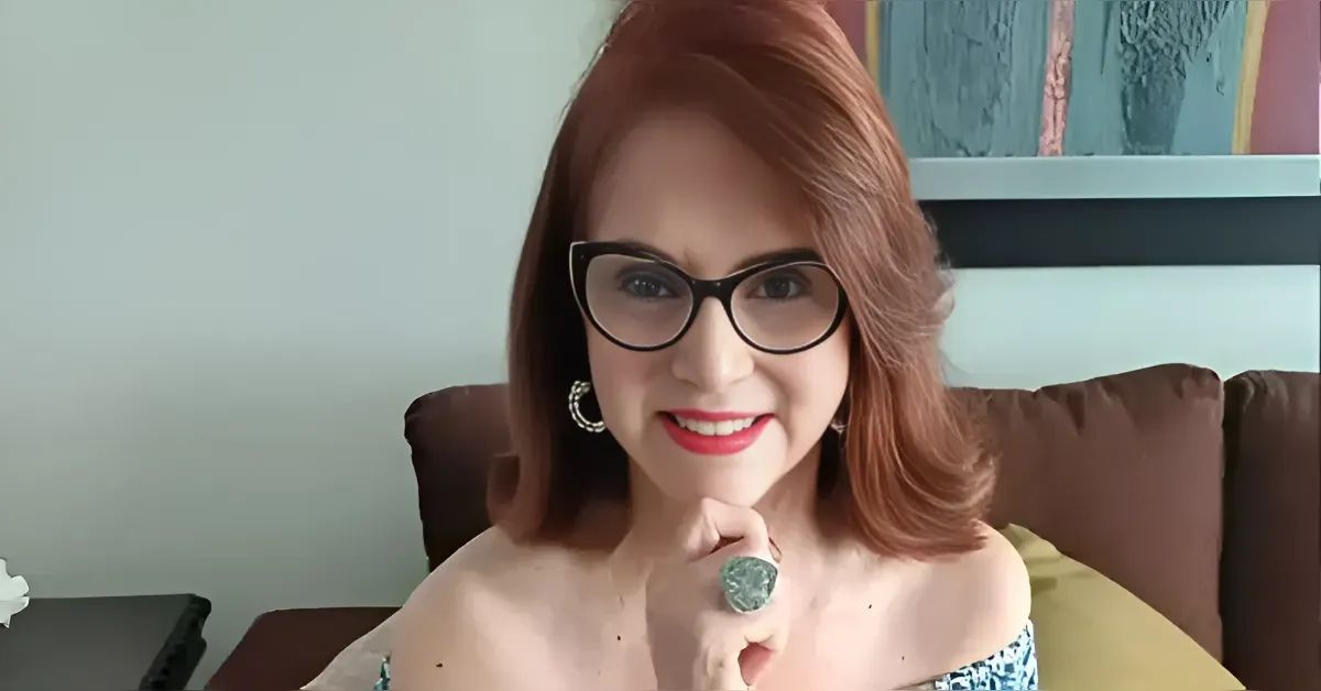 Smiling woman with red hair and black glasses sits on a brown couch, hand under chin showing a large turquoise ring.