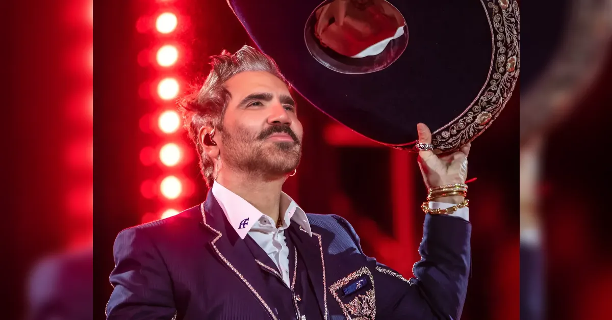 Male performer in a navy blazer with gold trim raises an ornate sombrero on stage under red lights.