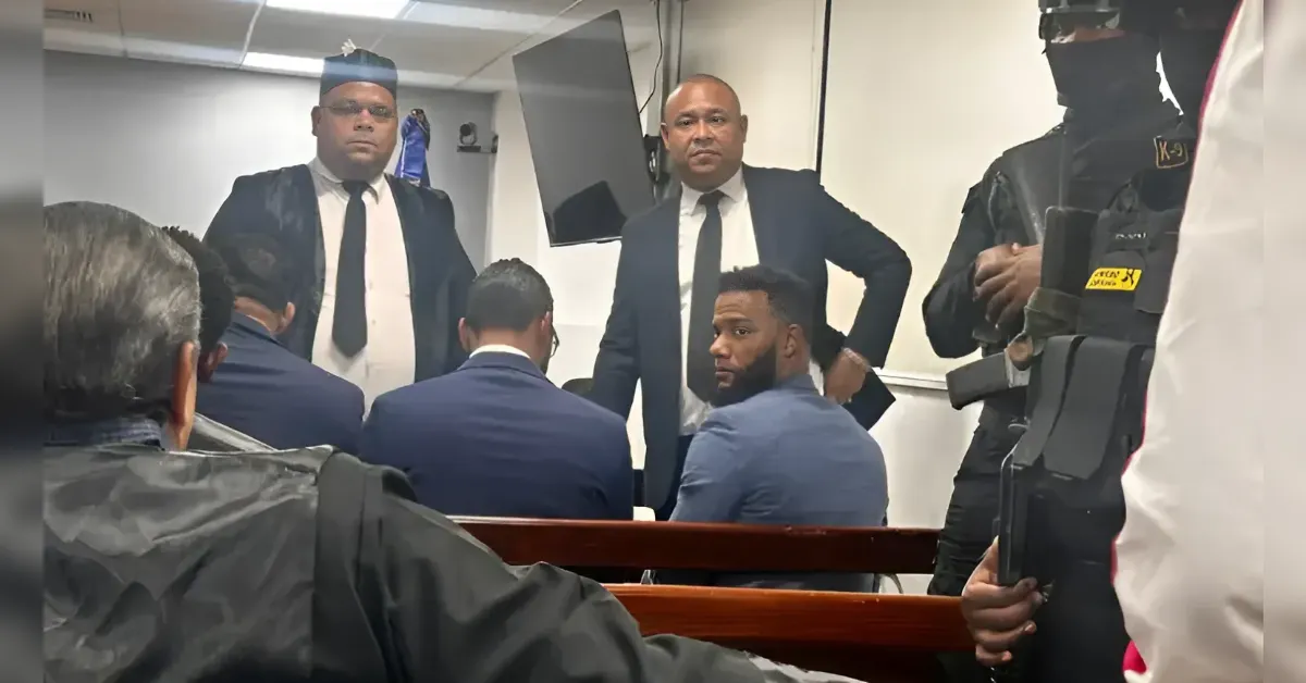 Group of men in suits seated in a courtroom-like room, with armed security officers standing nearby.