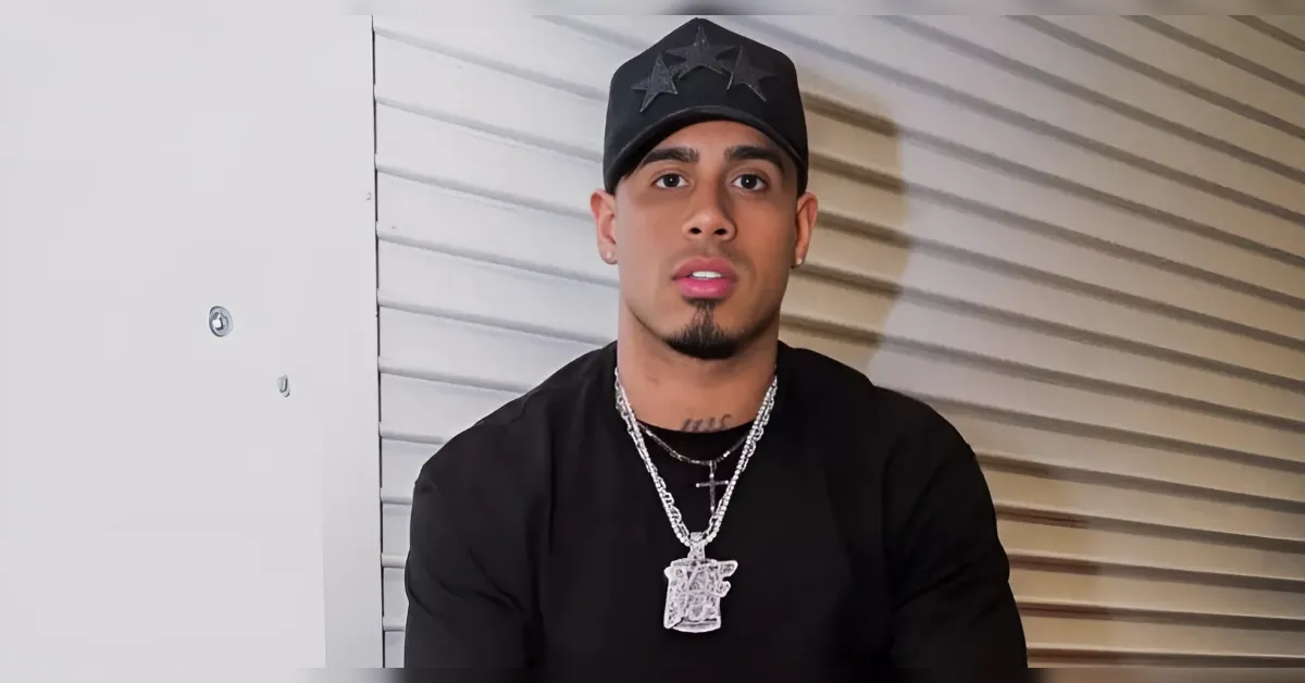 Portrait of a man in a black shirt and cap with star designs, wearing layered silver chains, posed against a striped wall.