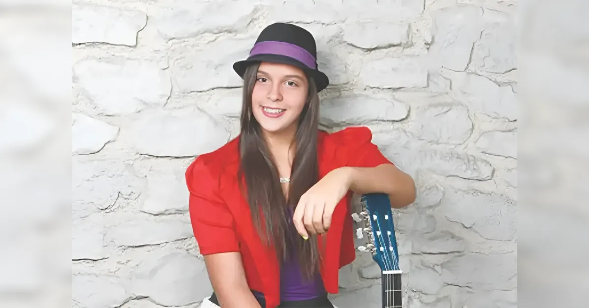 Young woman in a red blazer and purple top sits against a white stone wall, resting her arm on a blue guitar.