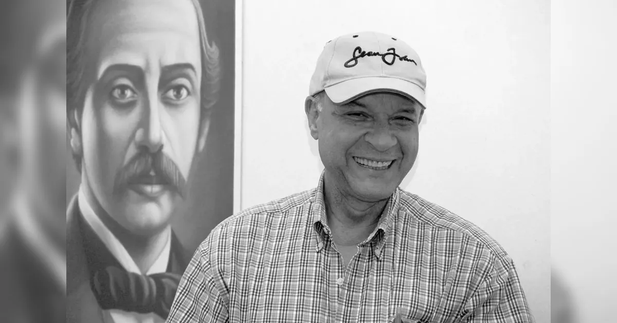 Smiling man in a plaid shirt and white cap standing beside a large black-and-white portrait of a man with a mustache on the wall.