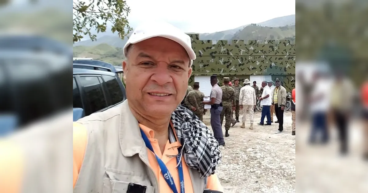 Man in a light vest and cap takes a selfie in front of a group of soldiers near a camouflaged tent in a mountainous outdoor area.