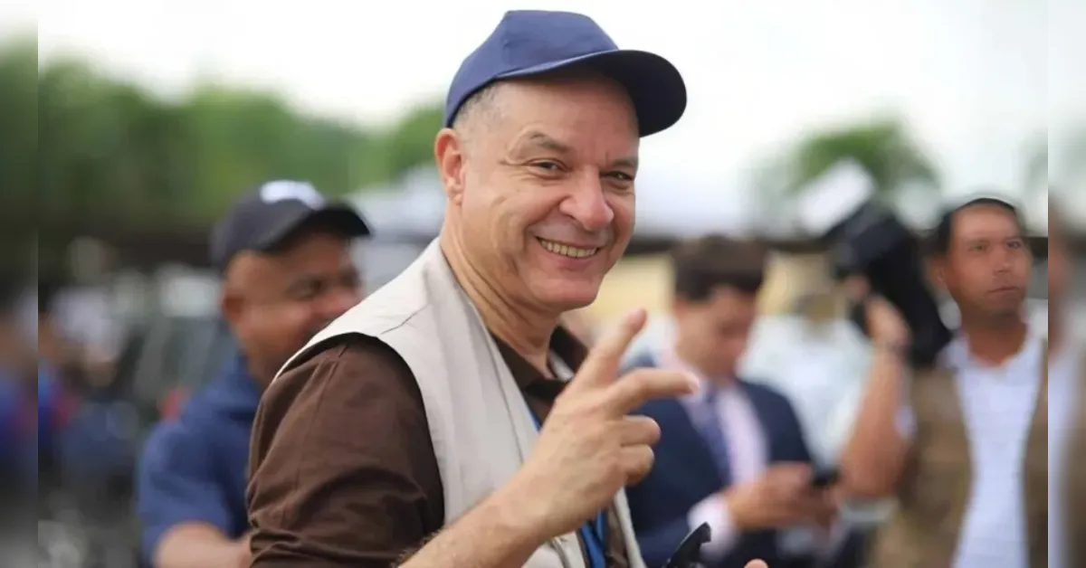 Smiling man in a blue cap making a peace sign at an outdoor event, with a blurred crowd in the background.