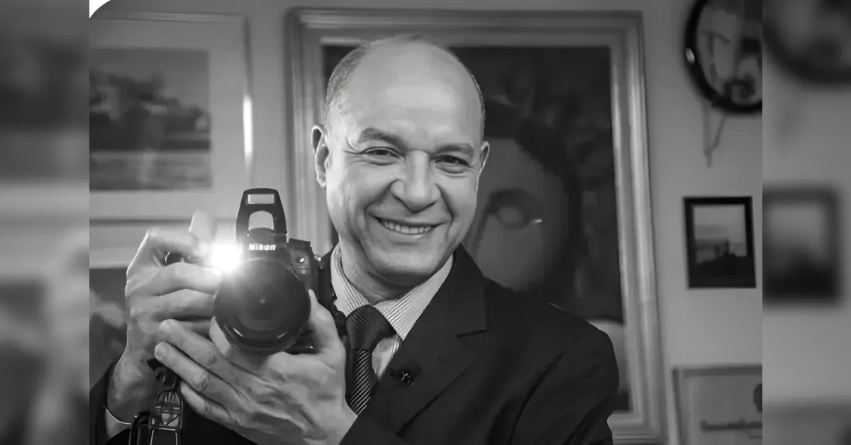 Smiling man in a suit holds a DSLR camera toward the viewer, flash glaring from the lens.