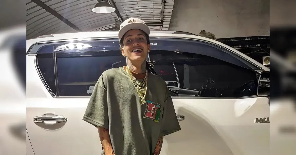 Person smiling in a loose green T‑shirt and white cap, posing beside a white SUV in a garage, wearing gold chains and earrings.