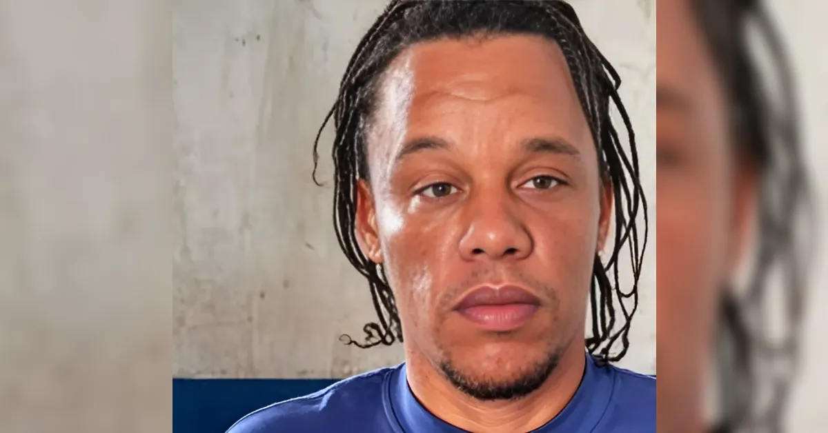 Close-up portrait of a man with dreadlocks and a blue shirt, looking at the camera against a plain wall background.