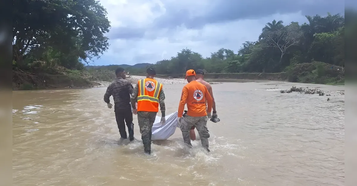 Rescuers wading in a muddy river carrying a person wrapped in a white sheet toward shore, wearing orange vests and camouflage gear, with a bare-chested companion nearby.