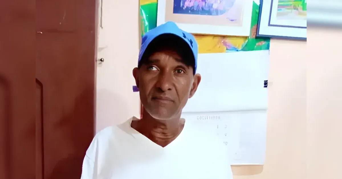 Portrait of a man wearing a blue cap and white shirt standing indoors near a door and framed art on the wall.