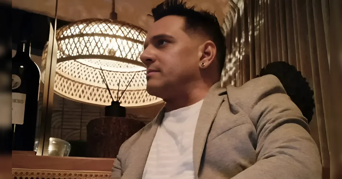Man in a light blazer and white shirt sits indoors beside a decorative lamp and wine bottle in a cozy setting, looking to the right.