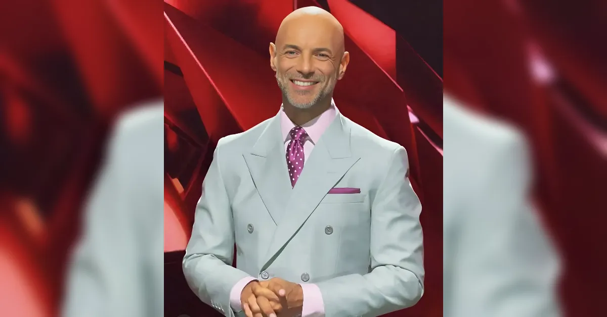 Smiling man in a light gray double-breasted suit, pink shirt, and purple polka-dot tie on a red abstract stage background.