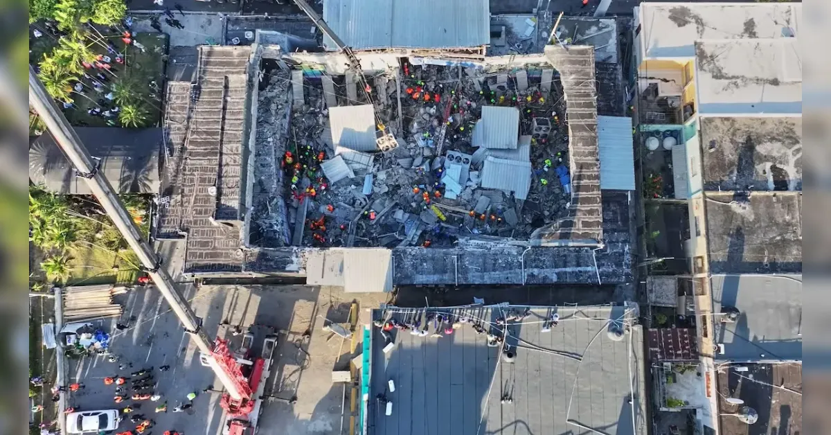 Aerial view of a collapsed building with rubble and numerous workers in orange vests around the site, with a crane overhead.