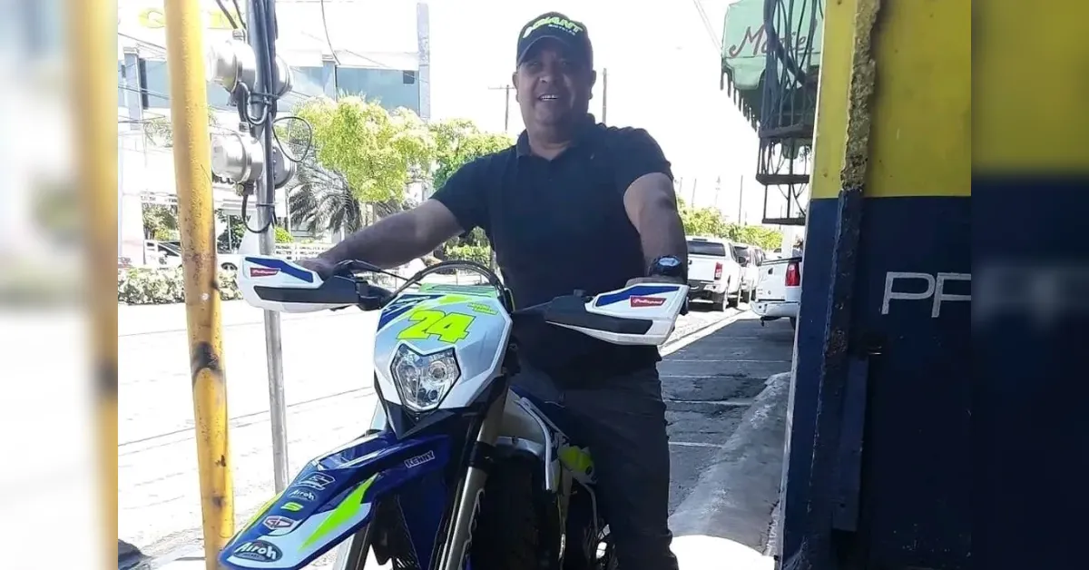 Man in a dark shirt and cap sits on a blue dirt bike with a neon yellow number 24, on a sunny street outside a shop.