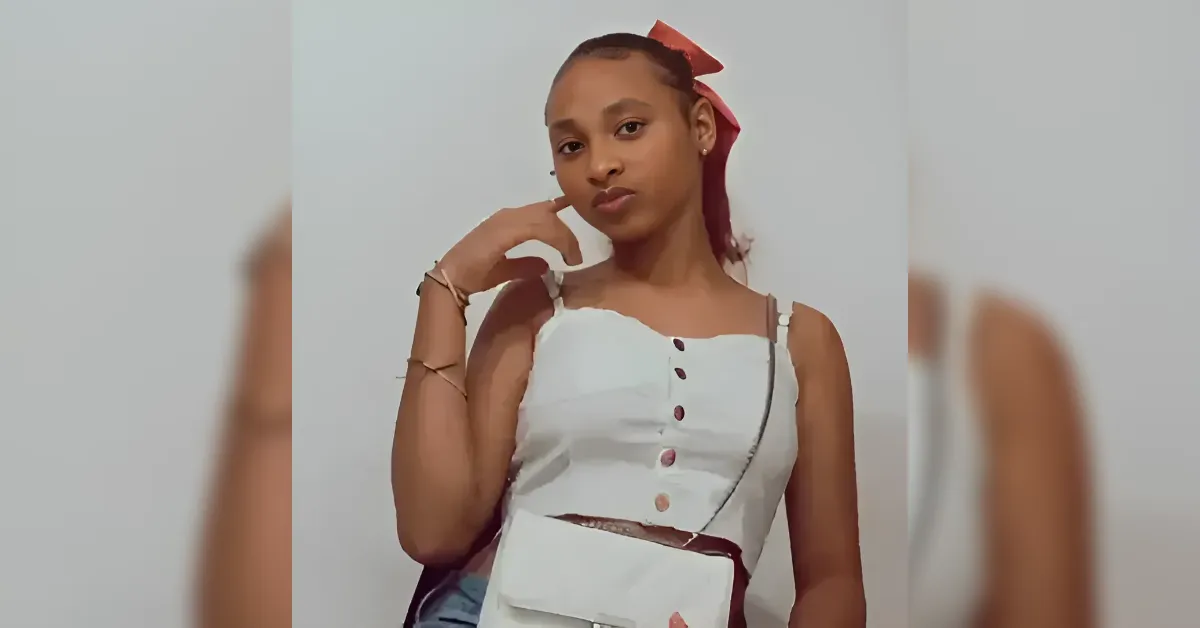 Young woman posing in a white sleeveless button-down top with a pink scarf in her hair and a crossbody bag.