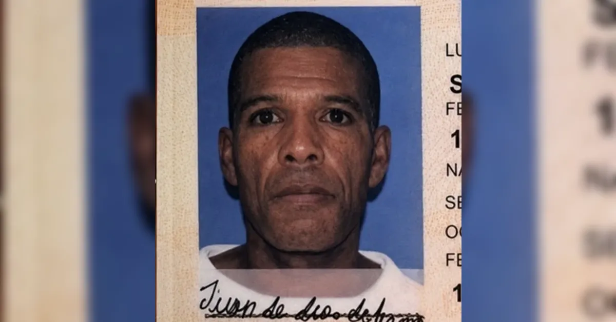 Mugshot photo of a man with short hair and a serious expression against a blue background, wearing a white shirt.