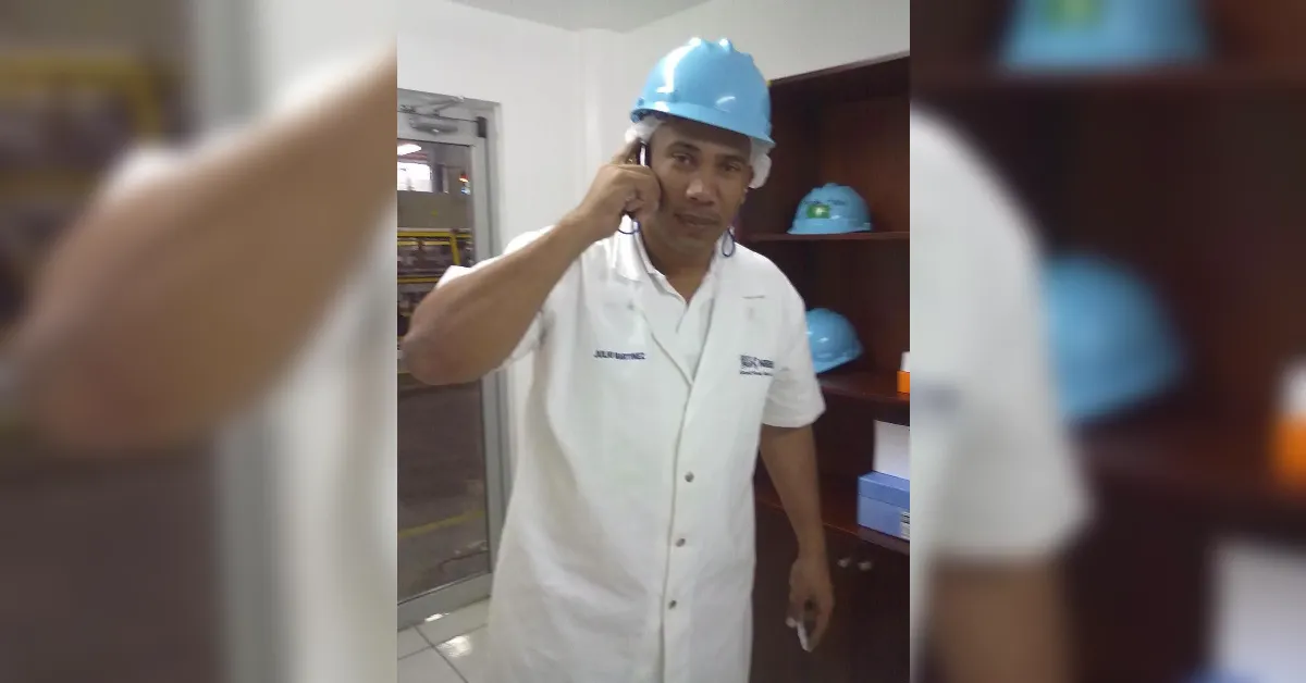 Man in a white lab coat and blue hard hat speaks on a mobile phone in an industrial room, with blue helmets on shelves behind him.