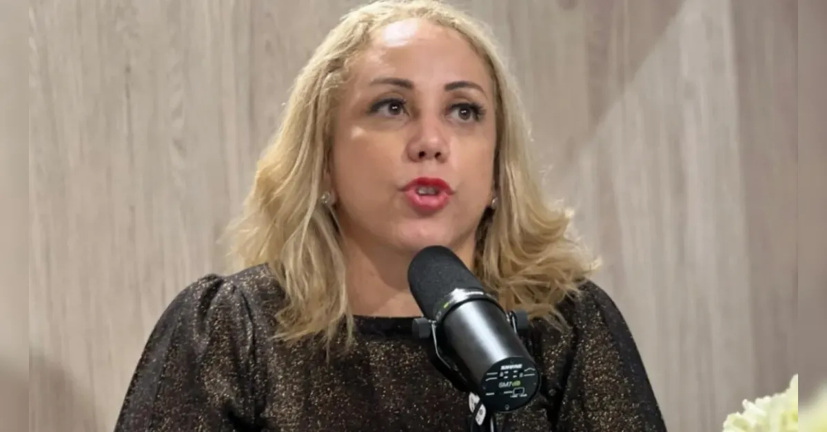 Woman with blonde hair, wearing a glittery black top, speaks into a microphone in a podcast or interview setting against a wood-paneled backdrop.