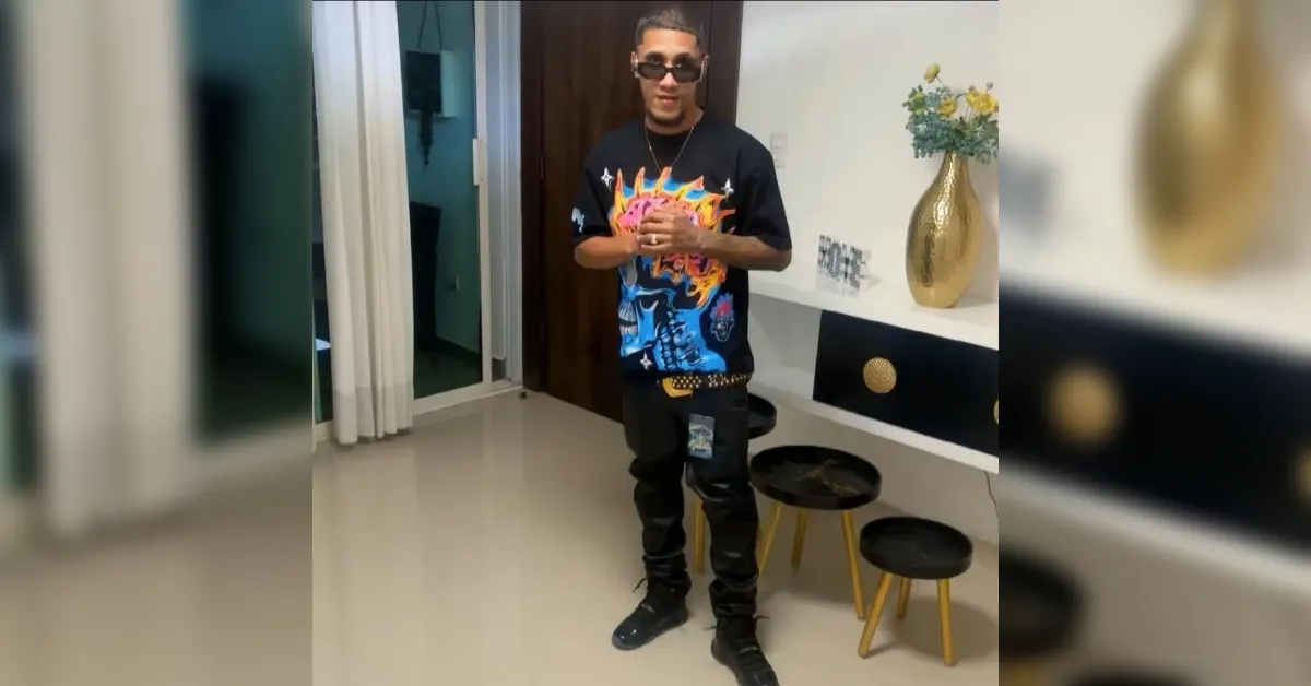 Man in a colorful graphic T-shirt and sunglasses stands with hands clasped indoors, modern white and black decor behind him.