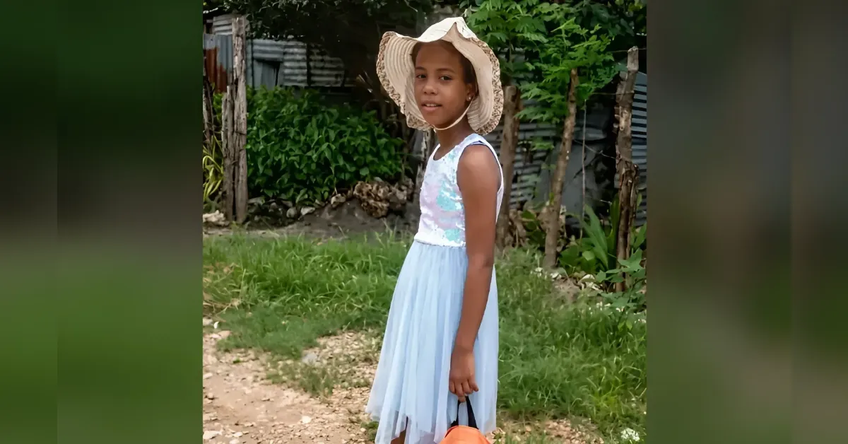 Young girl in a pale blue dress and wide-brimmed hat standing on a dirt path in a garden, holding an orange bag.