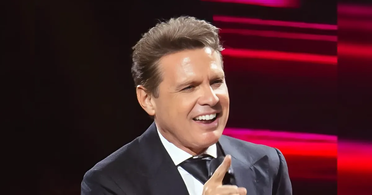 Man in a dark suit on a stage, smiling and gesturing with a raised index finger as he speaks or jokes around,