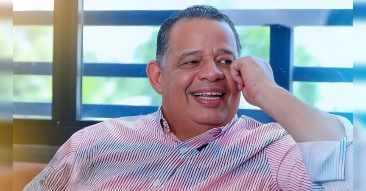 Smiling man in a pink striped shirt sits indoors, hand resting on his cheek.
