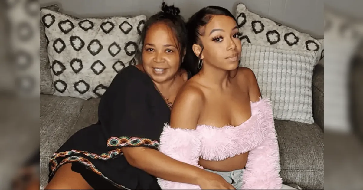 Two women sit close on a gray sofa, smiling; one wears a black dress with colorful trim, the other a pink fluffy off‑the‑shoulder top.